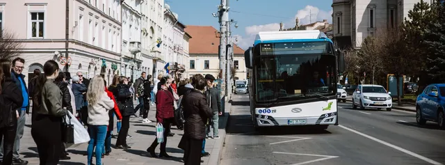 Emberek várakoznak egy belvárosi buszmegállóban, miközben egy fehér-kék városi busz áll meg mellettük egy régi épületekkel szegélyezett utcán, a forgalomban több személygépkocsi is látható.