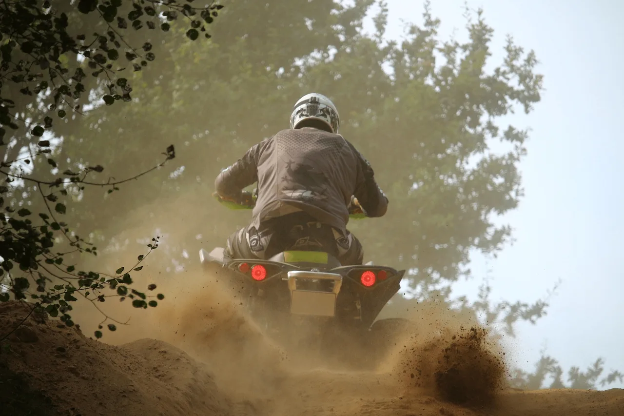 Hátulról fotózott négykerekű motoros (ATV) sisakos vezetője földúton, ahogy port és sarat felverve halad felfelé egy dombon, körülötte fák.
