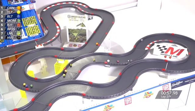 Golyós versenypálya (Marble Race) látható felülnézetből, kanyargós szürke pályával, amelyen színes márványgolyók gördülnek végig, mellette pontverseny táblázat és kör-visszaszámláló látható a képernyőn.