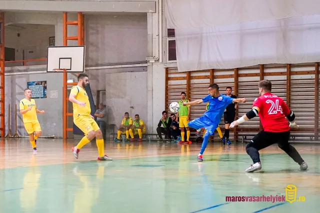 Teremlabdarúgó-mérkőzés sportcsarnokban: kék mezes játékos lövést végez a piros mezes, 24-es számú kapus felé, mögöttük sárga mezes csapattársak és tartalék játékosok figyelik a padról.
