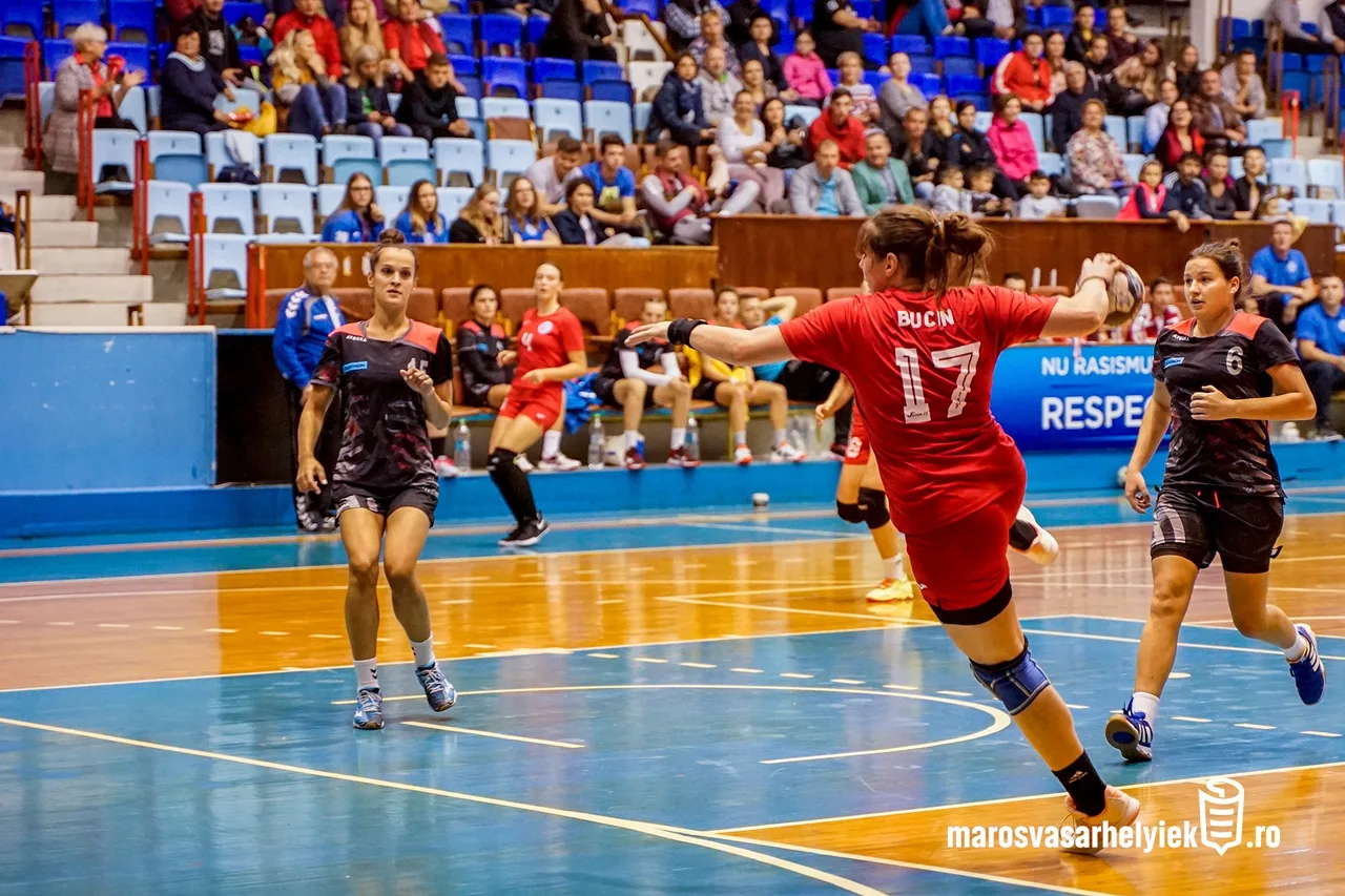 Női kézilabdamérkőzés egy sportcsarnokban: piros mezes, 17-es számú játékos (Bucin) felugorva dob kaput, mögötte fekete-piros mezes ellenfelek próbálják védekezni, a lelátón nézők ülnek.