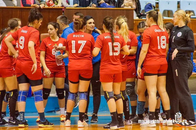 Piros mezes női röplabdacsapat áll körben egy időkérés vagy szünet alatt a sportcsarnokban, mellettük fekete öltözékű edző beszél hozzájuk, jobb oldalt egy fekete melegítős csapattag figyel.