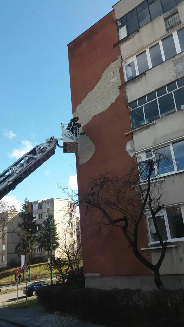 Tűzoltók emelőkosárban egy panelház vörös homlokzatán dolgoznak, ahol a vakolat egy része leomlott. A háttérben más tömbházak, fenyőfák és lombtalan fák láthatók tiszta, kék égbolt alatt.