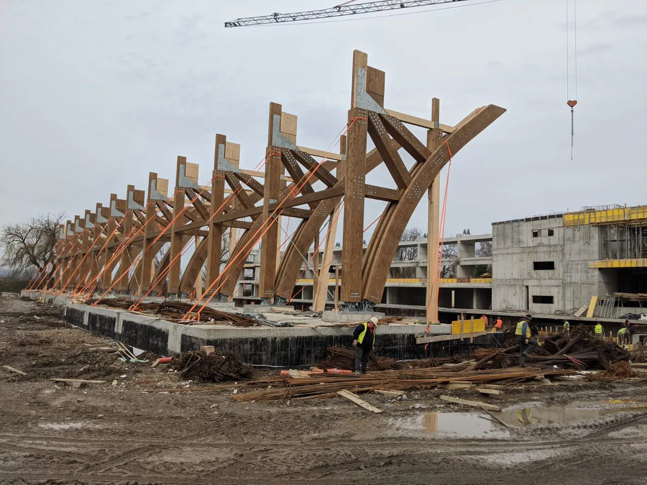 Építési területen fából készült ívelt tetőszerkezeti elemek sorakoznak egy betonalapon, mögöttük többszintes betonvázas épület látható, előtérben munkások, vasrudak és törmelék, felül daru.