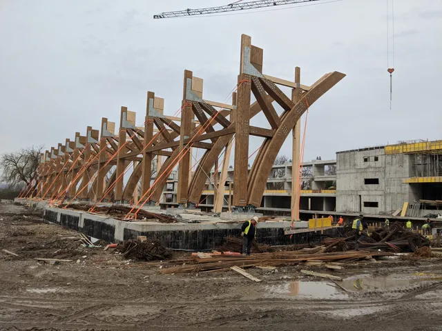 Építési területen fából készült ívelt tetőszerkezeti elemek sorakoznak egy betonalapon, mögöttük többszintes betonvázas épület látható, előtérben munkások, vasrudak és törmelék, felül daru.