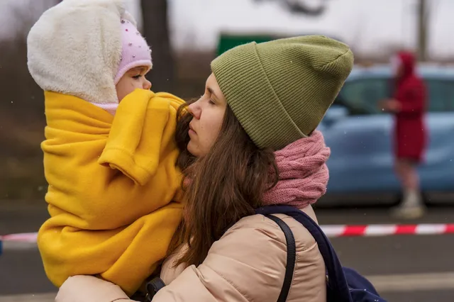 Egy zöld sapkás és rózsaszín sálat viselő nő karján tart egy sárga kabátba, fehér szőrmés kapucnis sapkába bújtatott kisgyermeket, mögöttük autó és útelzáró szalag látszik egy utcán.
