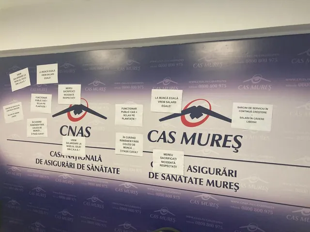 A CNAS és CAS Mureș (Maros megyei Egészségbiztosítási Pénztár) logóival díszített fal, amelyre kézzel írt papírlapokat ragasztottak fel, panaszos-tiltakozó jellegű szövegekkel a munkakörülményekről és a bérekről.