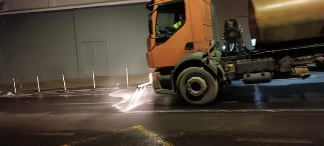 Éjszaka egy narancssárga utcamosó teherautó permetezi a vizet az útra, sofőrje a fülkében látható; a háttérben szürke fal és útelválasztó oszlopok láthatók.