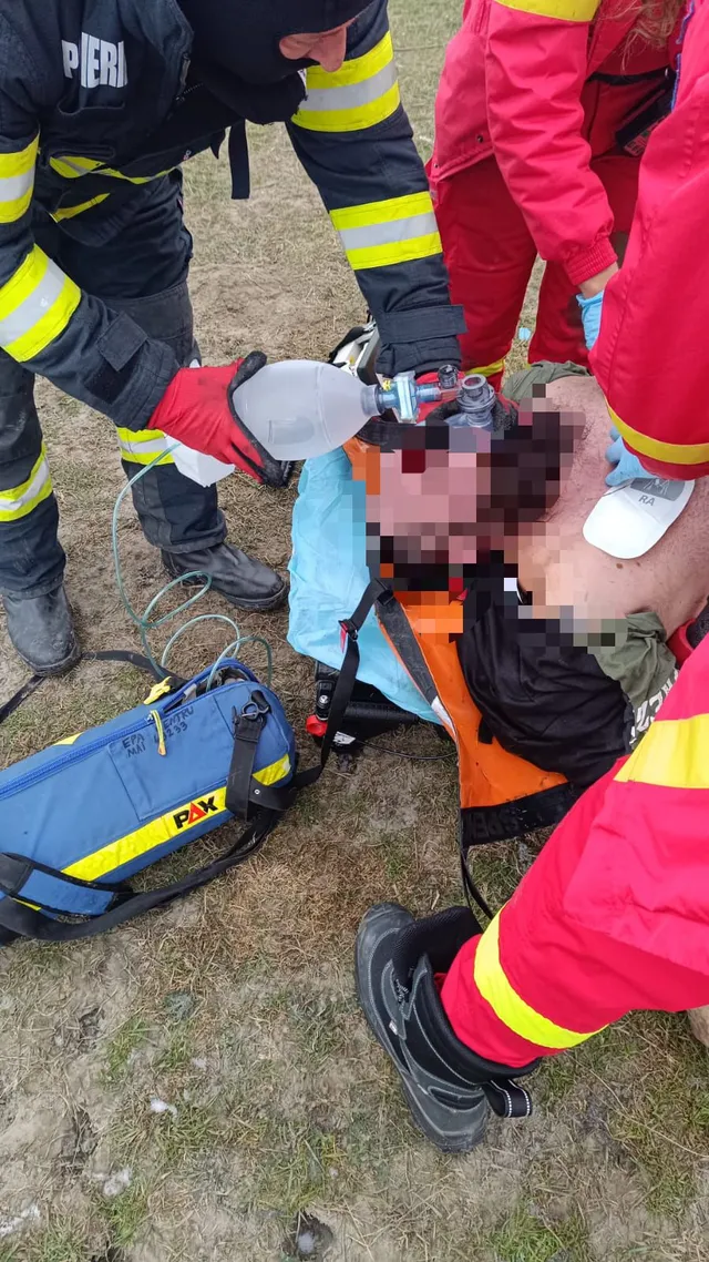 Fekvő sérült férfit ápolnak a földön mentőalakok: egy tűzoltó lélegeztetőballont tart az arcára, mellette mentős szakemberek pirosnarukú ruhában láthatók, valamint egy kék mentőtáska van a fűben.
