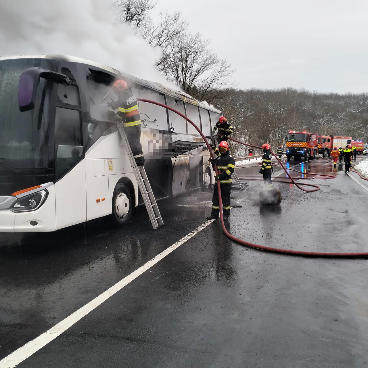 Kigyulladt távolsági autóbusz oltása egy útszakaszon, tűzoltók létrán és tömlőkkel dolgoznak, a busz oldalából és tetejéből sűrű füst gomolyog, háttérben tűzoltóautók sorakoznak.