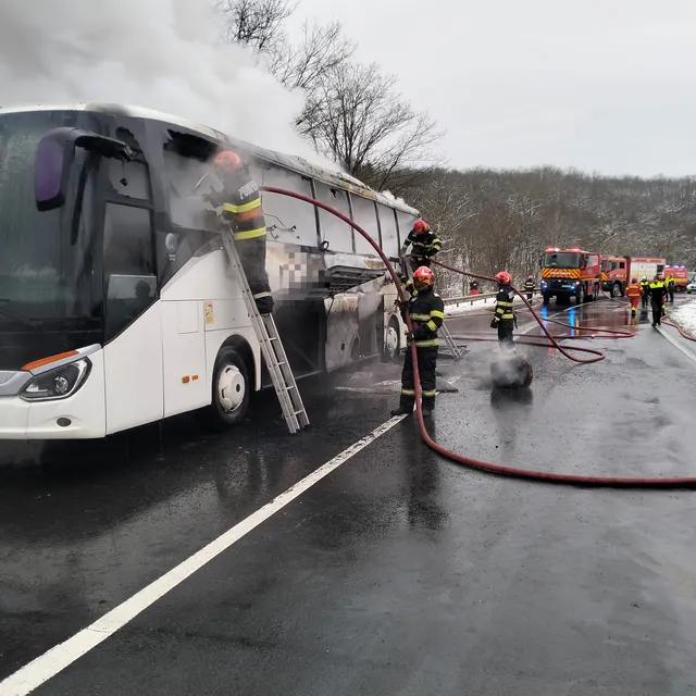 Kigyulladt távolsági autóbusz oltása egy útszakaszon, tűzoltók létrán és tömlőkkel dolgoznak, a busz oldalából és tetejéből sűrű füst gomolyog, háttérben tűzoltóautók sorakoznak.
