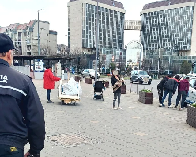 Városi téren rendőr áll előtérben egy csoport háttal, mögötte egy nő gitározik, valamint emberek húznak kézikocsit és babakocsit, körülöttük üzletházak és forgalom.