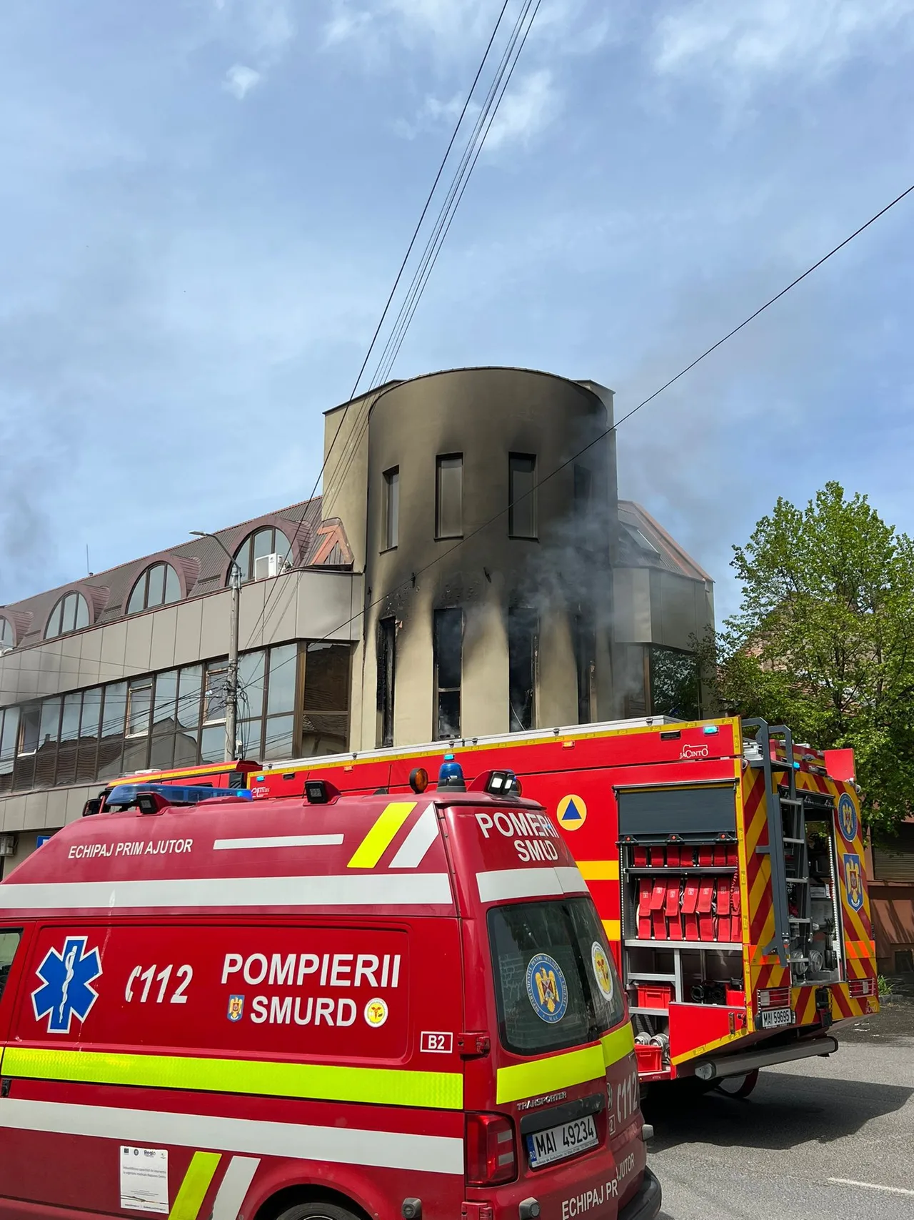 Egy épület felső szintjének ablakai és homlokzata kormosan, füstösen látszanak tűz után, előtte SMURD és pompierii feliratú tűzoltóautók parkolnak az utcán.
