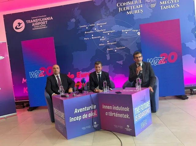 Három férfi ül fotelekben egy sajtótájékoztatón, középen és jobb oldalon mikrofonokba beszélnek. A háttérben a Transylvania Airport és a Wizz Air 20. évfordulóját hirdető molinó látható európai úticélokat összekötő térképpel, előttük Wizz Air feliratú kockák.
