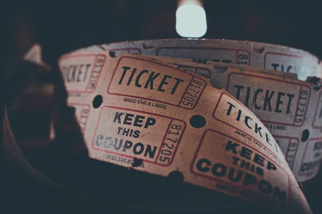 Közelkép sorszámozott „TICKET” feliratú belépőjegy-tekercsekről, amelyeken „Keep This Coupon” felirat is olvasható, háttérben elmosódott fényforrással.
