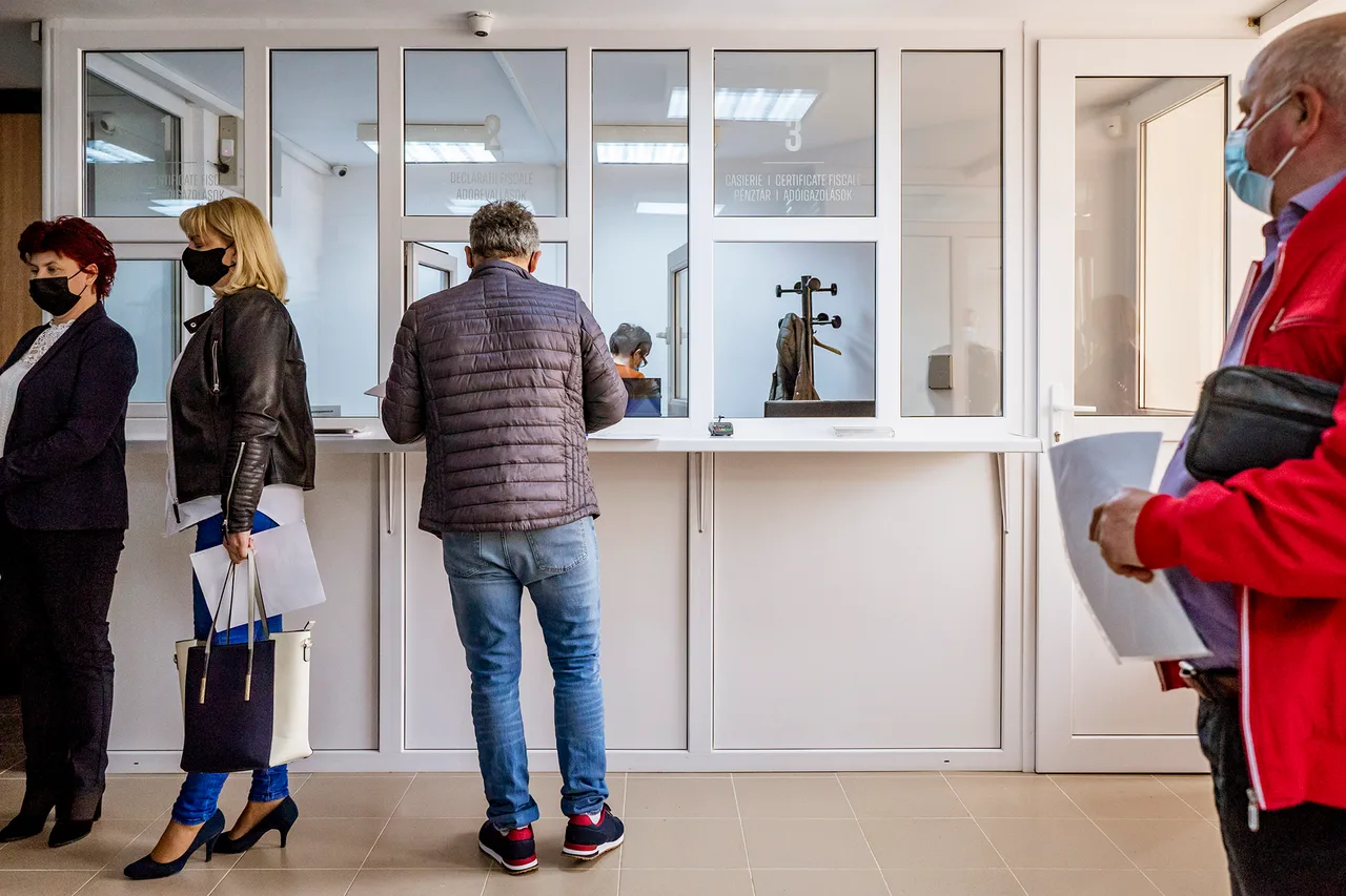 Emberek sorban állnak egy hivatal ügyfélszolgálati ablakainál, arcmaszkot viselnek, dokumentumokat tartanak kezükben, miközben az egyik pénztárablaknál ügyintézés folyik.