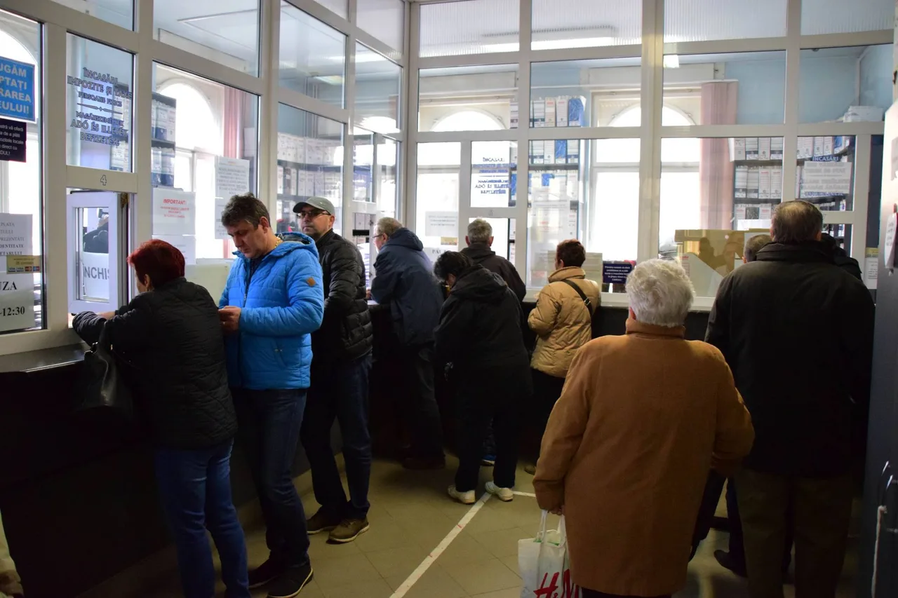 Emberek sorban állnak egy adóhivatal ügyfélfogadó termében, ablakos pénztárablakoknál intézik ügyeiket, kabátban, mivel beltéri, de hűvös helyszín.