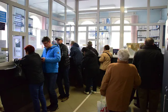 Emberek sorban állnak egy adóhivatal ügyfélfogadó termében, ablakos pénztárablakoknál intézik ügyeiket, kabátban, mivel beltéri, de hűvös helyszín.