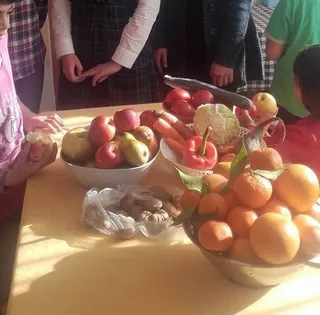 Sárga asztalon tálak és zacskók zöldségekkel és gyümölcsökkel: alma, körte, paradicsom, sárgarépa, karfiol, mandarin, narancs, gyömbér. Az asztal körül gyerekek állnak, arcuk nem látszik a képen.