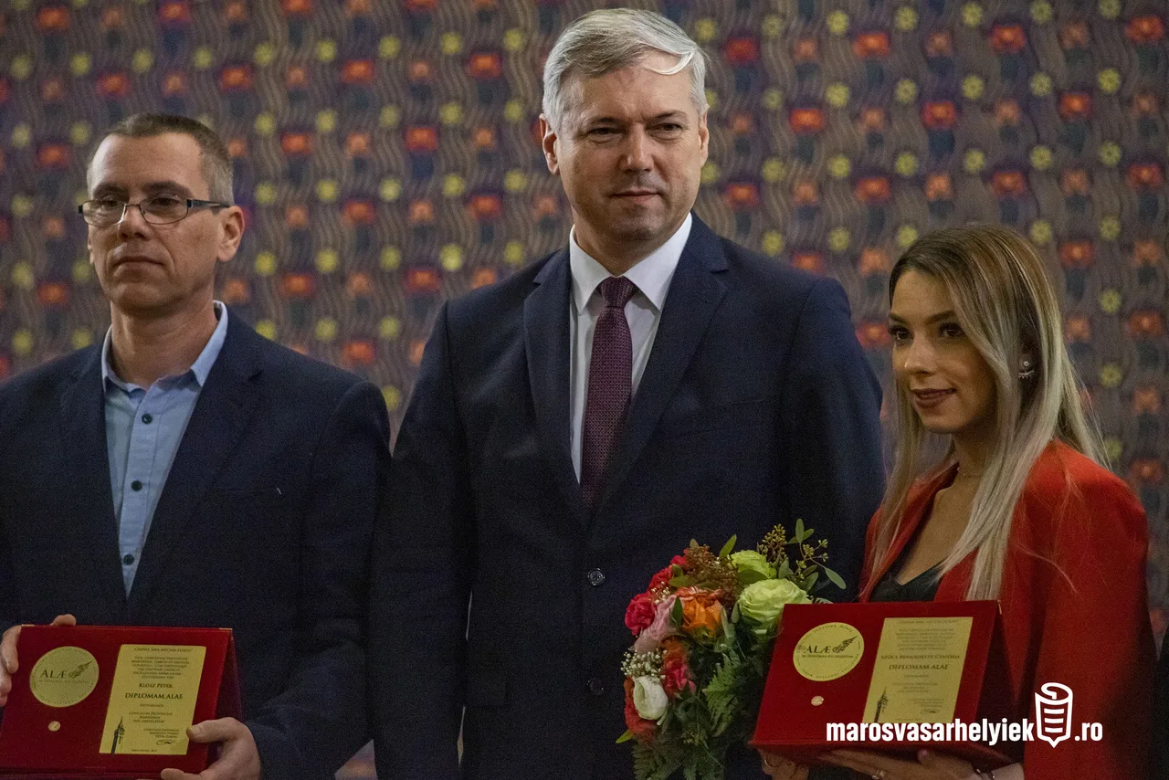 Két férfi és egy nő állnak egymás mellett díszes háttér előtt, a bal oldali férfi és a jobb oldali nő piros díszdobozban lévő okleveleket tartanak a kezükben, a nő mellett egy virágcsokor is látható.