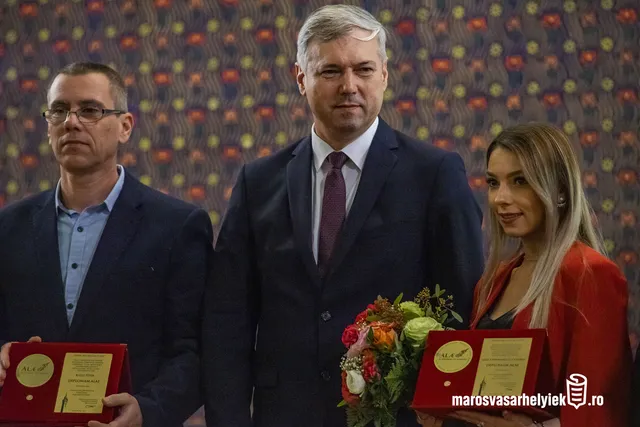 Két férfi és egy nő állnak egymás mellett díszes háttér előtt, a bal oldali férfi és a jobb oldali nő piros díszdobozban lévő okleveleket tartanak a kezükben, a nő mellett egy virágcsokor is látható.