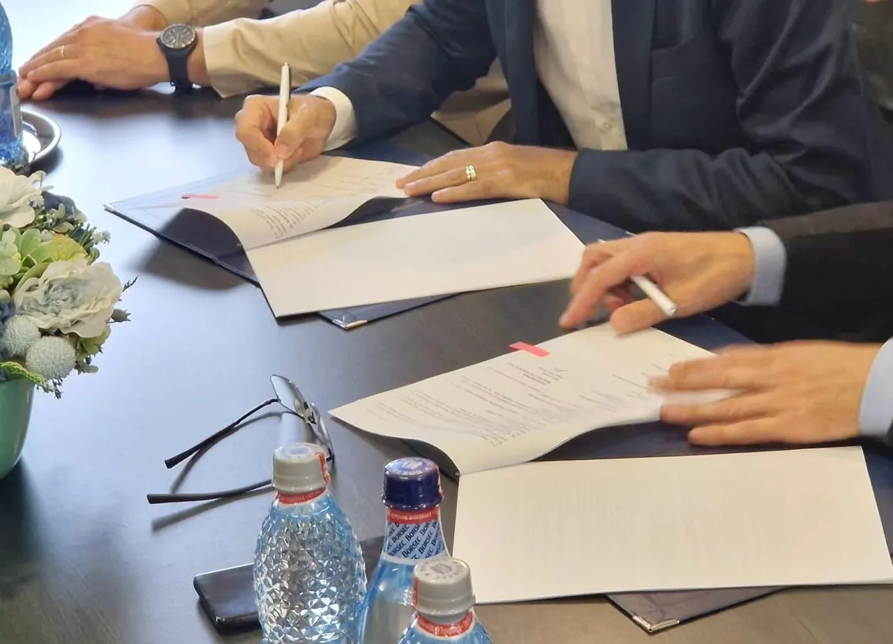 Öltönyös emberek asztalnál ülve papírokat írnak alá, előttük dossziék, szemüveg és ásványvizes üvegek, mellettük egy virágdísz.