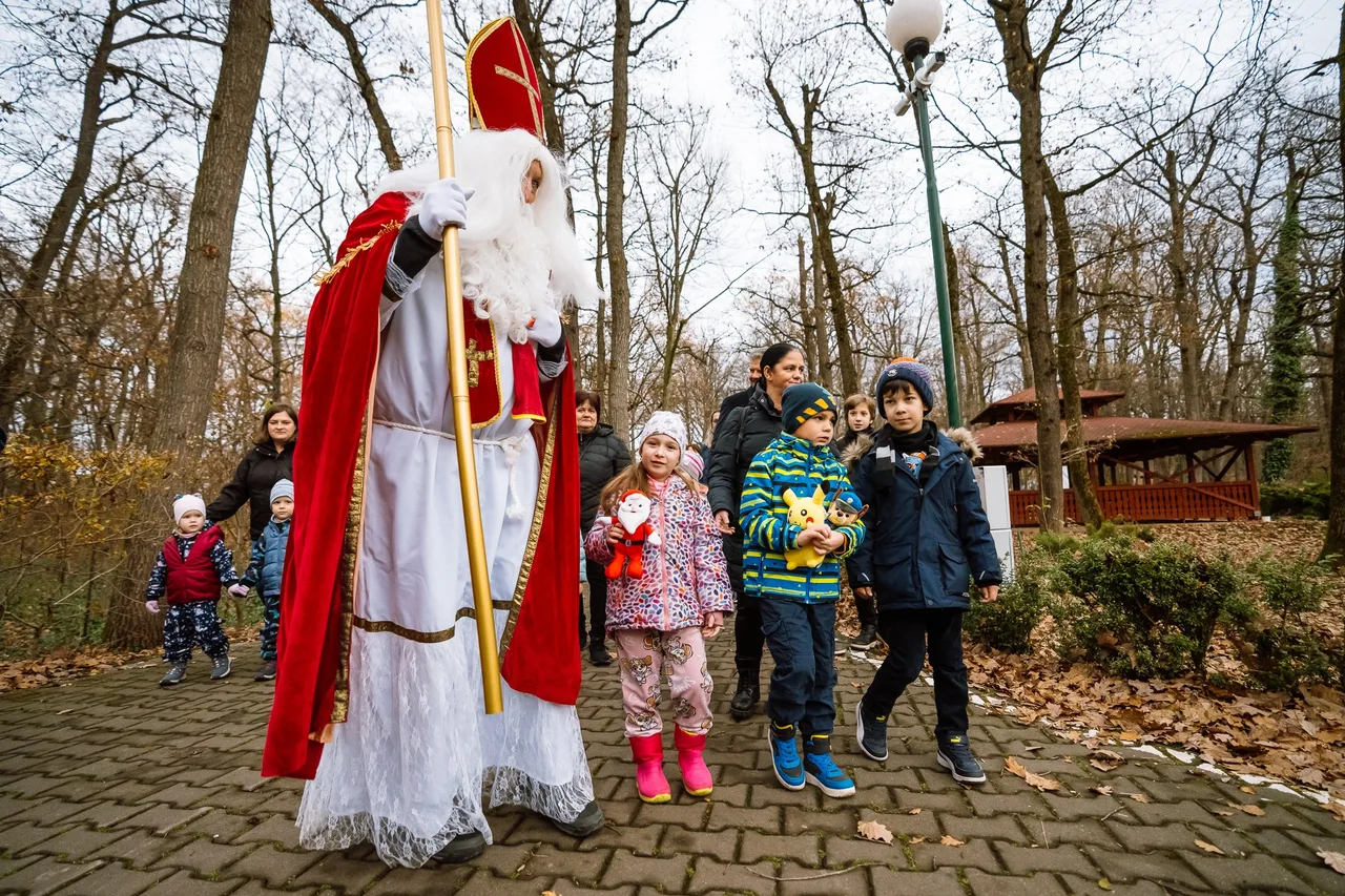 Püspöki (mitrás) öltözetű Mikulás-alakítás sétál egy parkos ösvényen, botot tart a kezében, körülötte gyerekek és felnőttek kísérik, egy kislány és egy kisfiú kis Mikulás- és rajzfilmfigurás plüssjátékokat tart a kezében.