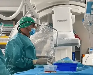Egy zöld sebészi köpenyt, sapkát és maszkot viselő orvos áll egy Philips márkájú angiográfiás (érképalkotó) berendezés mellett a kórházi műtőben, kezében sebészi eszközzel dolgozik egy steril kék terítővel letakart eszközasztalnál.
