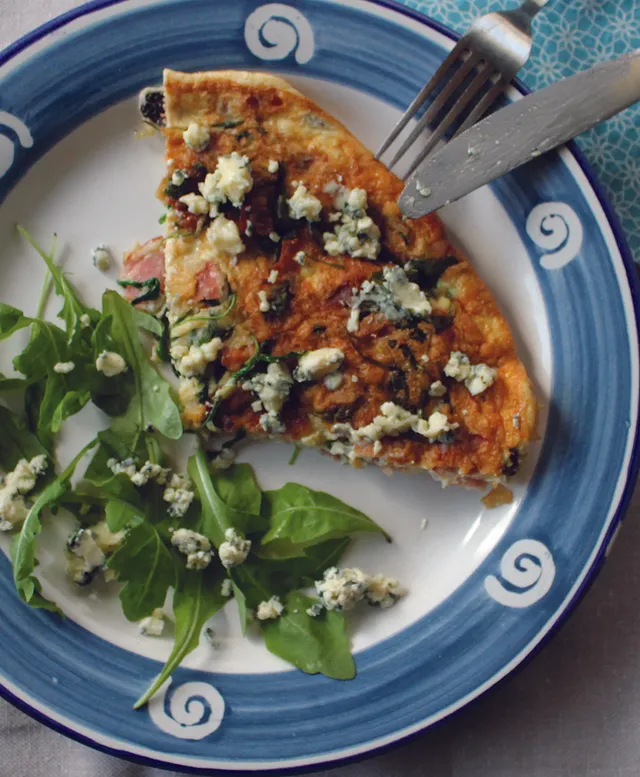 Kék-fehér mintás tányéron egy szelet sajtos-zöldséges quiche vagy omlett, mellette rukkolasaláta kék sajt morzsával megszórva, a tányéron villa és kés látható.