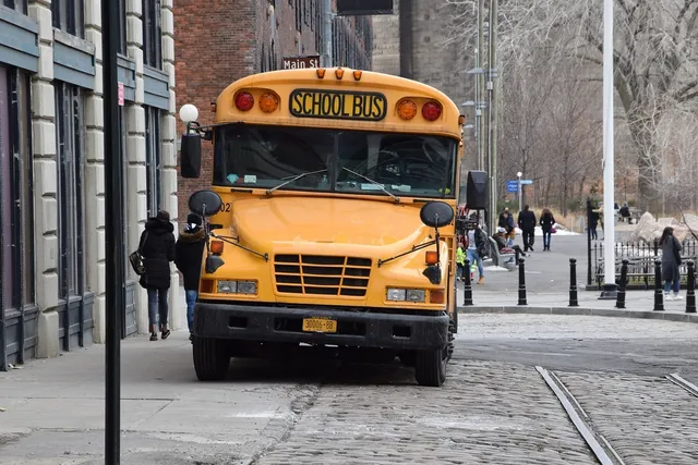 Sárga amerikai iskolabusz áll egy macskaköves utcán, előtte kockakő járda, mellette a Main St. tábla látható; a bal oldali járdán két gyalogos halad el a busz mellett, a háttérben téglaépületek és néhány további járókelő látszik.