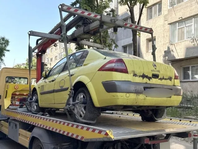 Sárga személygépkocsi van felpakolva egy autómentő (vontató) platójára, mögötte panelházak és fák láthatók egy városi utcában.