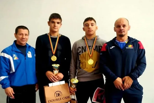 Négy férfi áll egymás mellett fehér háttér előtt: két edző sportfelszerelésben szélről, középen két fiatal sportoló érmekkel a nyakukban, kupákkal és ajándéktáskával a kezükben.