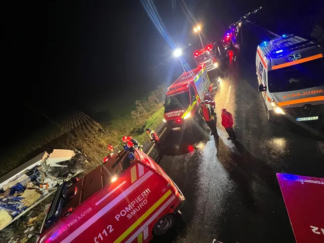 Éjszakai közúti baleset helyszíne esőben: tűzoltó- és mentőautók, kék és piros villogó fényekkel, az út szélén szétroncsolódott jármű törmelékei, több egyenruhás beavatkozó áll az úton.