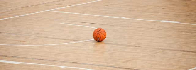 Egy narancssárga kosárlabda hever egy fényes parkettás sportcsarnok padlóján, a pálya fehér vonalai láthatók körülötte.
