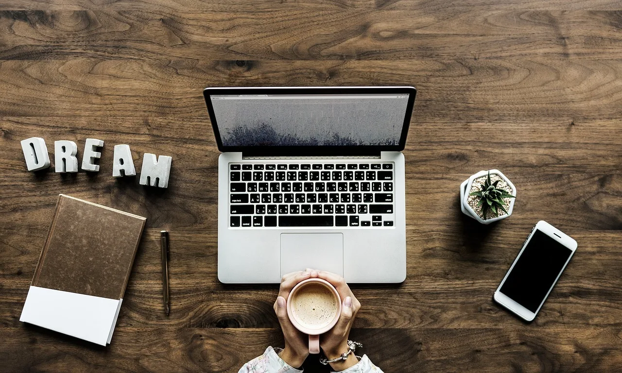 Felülnézeti kép egy fa asztalról: laptop, mellette egy csésze kávét tartó két kéz, notebook, toll, DREAM feliratú betűdíszek, egy kis kaktusz cserépben és egy okostelefon.