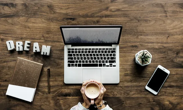 Felülnézeti kép egy fa asztalról: laptop, mellette egy csésze kávét tartó két kéz, notebook, toll, DREAM feliratú betűdíszek, egy kis kaktusz cserépben és egy okostelefon.