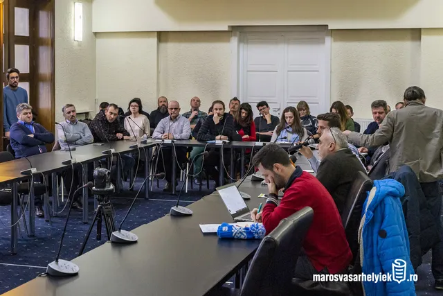 Tanácsterem asztalok köré ülve, mikrofonokkal felszerelt asztaloknál mintegy két tucat ember vesz részt egy nyilvános vitán, néhányan mikrofonba beszélnek, mások jegyzetelnek vagy hallgatnak.