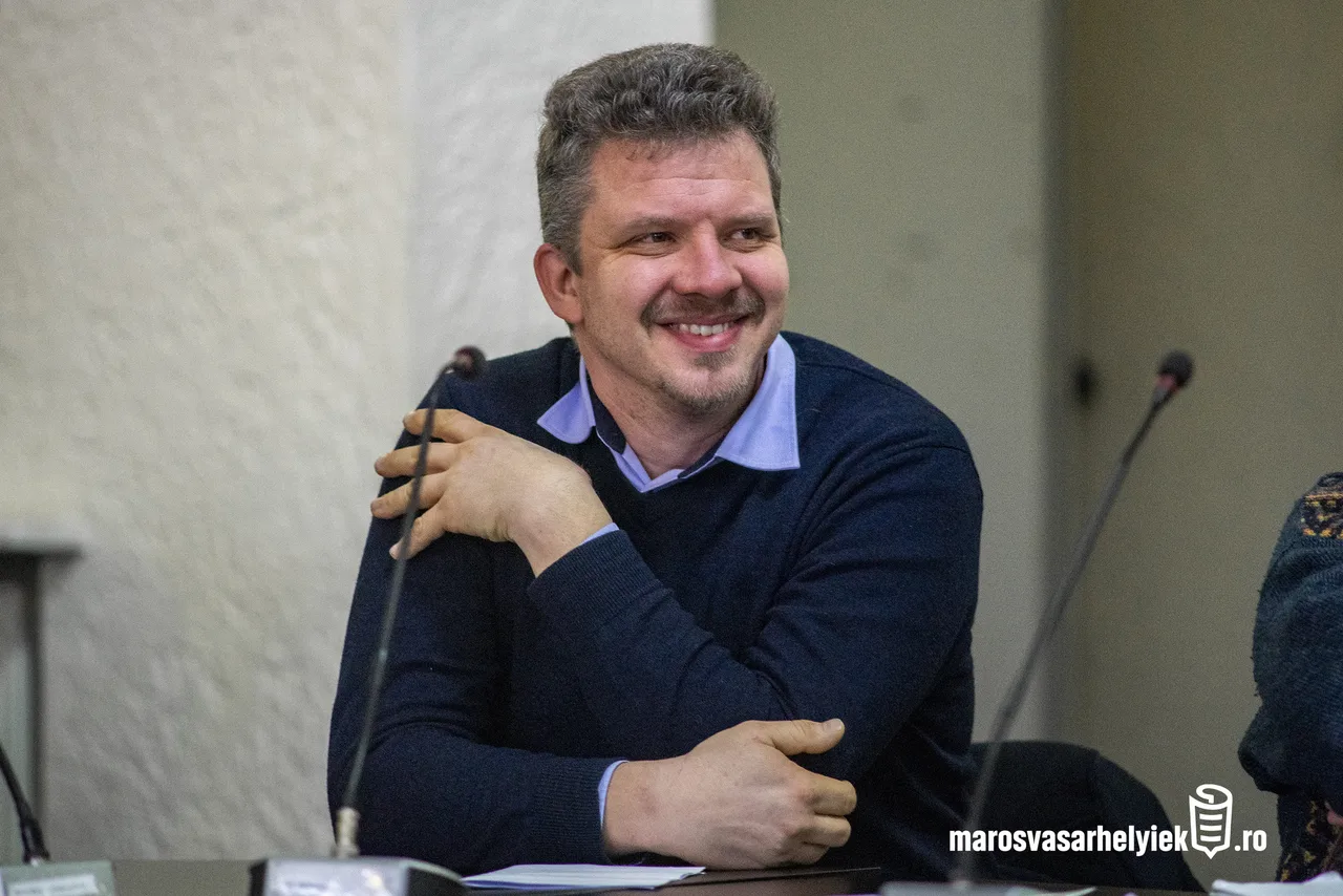 Sötétkék pulóvert és világoskék inget viselő férfi mosolyogva ül egy asztalnál, mikrofonok előtt, tanácskozóterem-szerű helyiségben.
