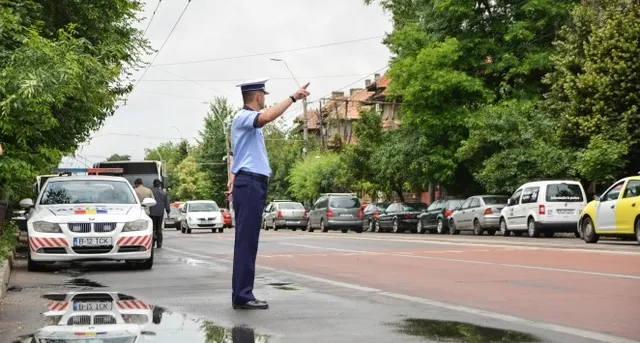 Rendőr egyenruhában áll az utcán, kézzel irányítja a forgalmat egy fás, parkoló autókkal szegélyezett úton, mellette rendőrségi autó áll.
