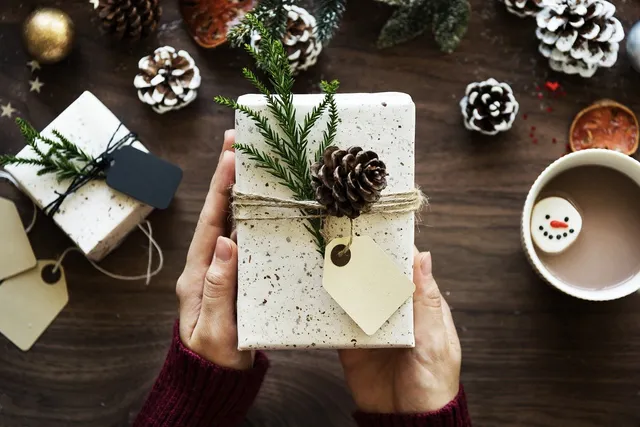 Két kéz tart egy karácsonyi csomagolópapírba tekert ajándékdobozt, amelyet zsinór, fenyőág, tobozdísz és papír ajándékcímke díszít. A háttérben faasztalon további csomagolt ajándékok, tobozok, fenyőágak, karácsonyi gömb és egy csésze forró csokoládé hócsata-keksszel láthatók.