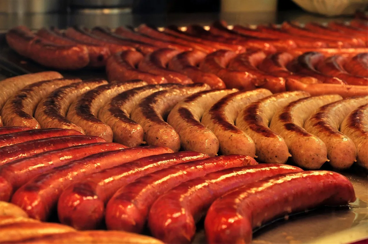 Sültkolbászok és virslik sorakoznak szorosan egymás mellett grillrácson, több különböző fajta, eltérő pirulási fokkal és színnel.