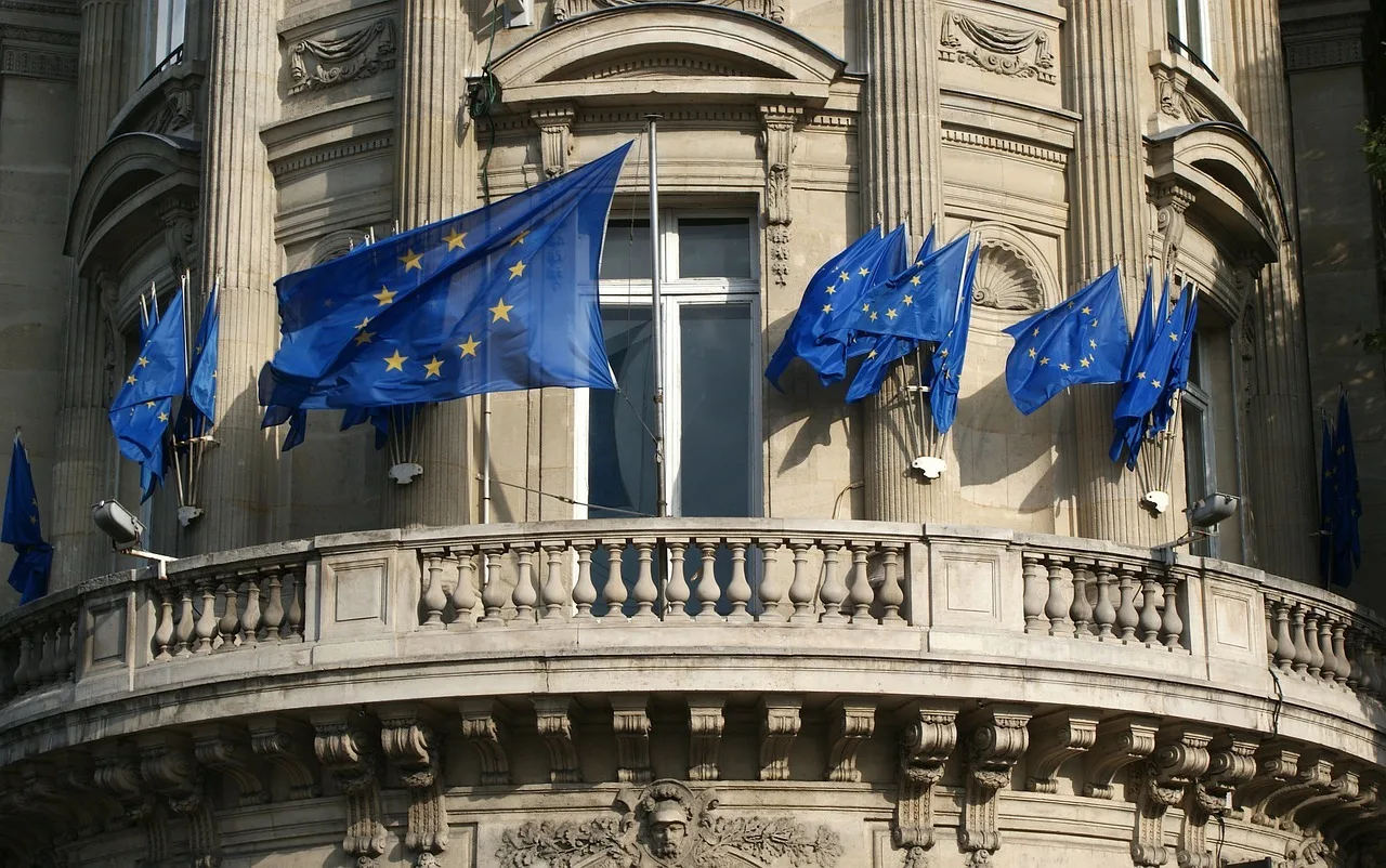 Egy klasszikus stílusú, díszes kőfaragású épület erkélyén és homlokzatán több európai uniós zászló lobog a szélben.