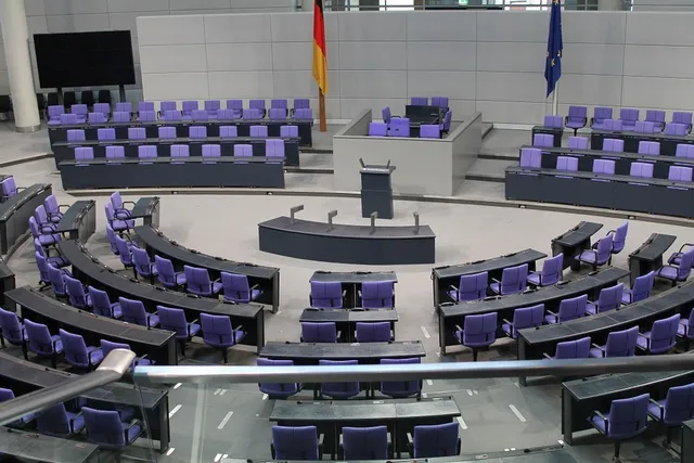 Üres parlamenti ülésterem lila színű ülésekkel, félkör alakban elrendezett padsorokkal, középen szónoki emelvénnyel, a háttérben német és európai uniós zászlóval.