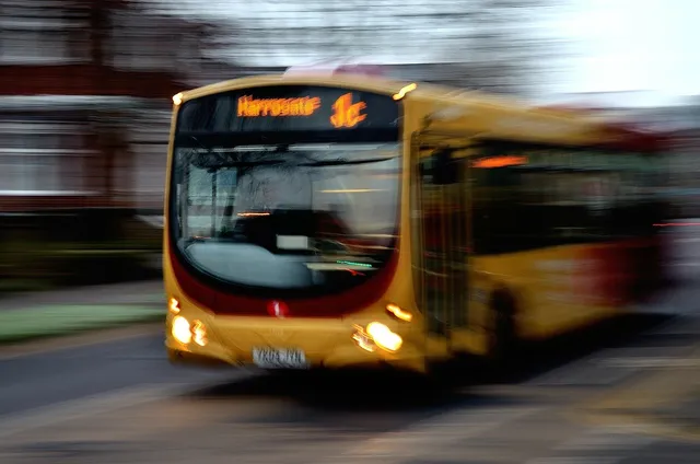 Sárga, csuklós városi autóbusz halad az úton, erőteljes mozgás-életlenséggel fényképezve; az elektronikus kijelzőn 'Harrogate 1c' felirat látszik.