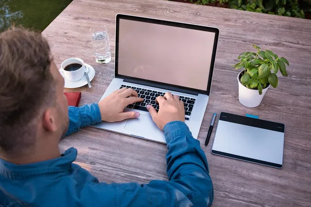 Egy férfi kültéri faasztalnál ül, kék dzsekiben, és laptopon gépel. Az asztalon kávé, pohár víz, egy cserepes bazsalikom és egy rajztábla tollal található.