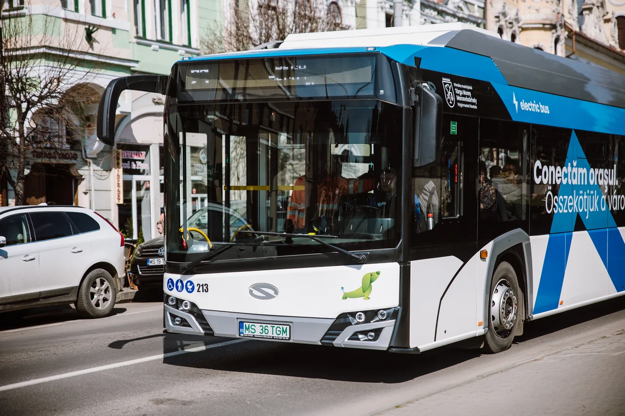 Fehér-kék elektromos autóbusz halad egy városi utcán, oldalán „Conectăm orașul / Összekötjük a várost” felirat és az önkormányzat logója, mellette parkoló autók és régi épületek láthatók.