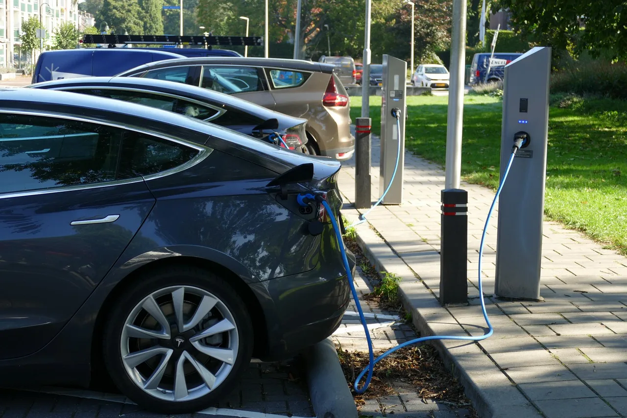 Egy parkolóban Tesla elektromos autó töltőkábellel csatlakoztatva egy oszlopos töltőállomáshoz, mögötte további parkoló autók és egy másik töltőoszlop látható.