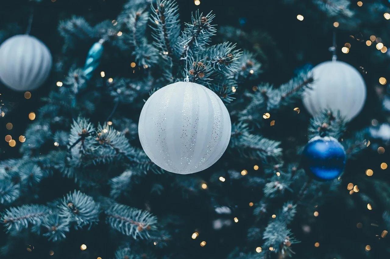 Közeli felvétel egy karácsonyfáról, melyen fehér és kék gömbdíszek lógnak, a háttérben elmosódott fényfüzér fényei látszanak.