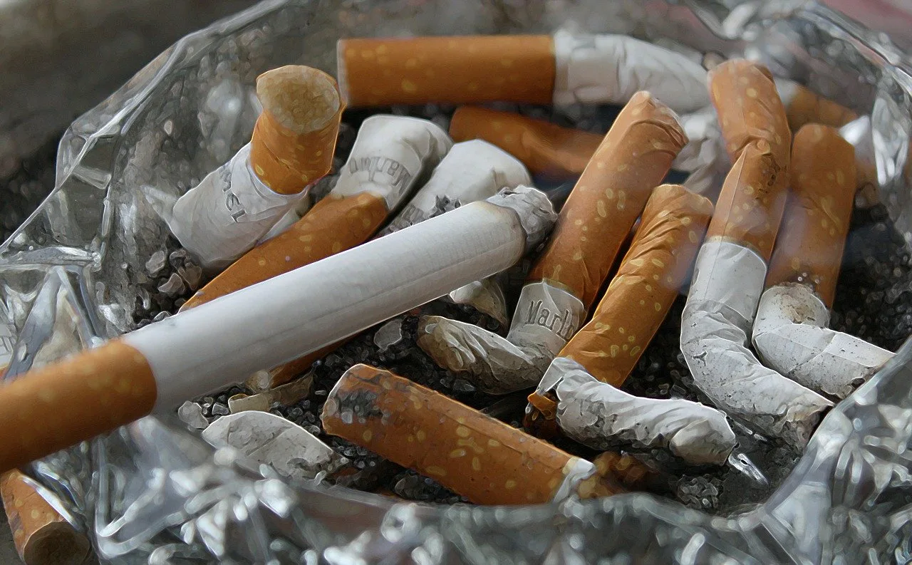 Közelről fotózott üvegtele hamutartó, tele elnyomott cigarettacsikkekkel és hamuval, elöl egy még égő cigarettával.
