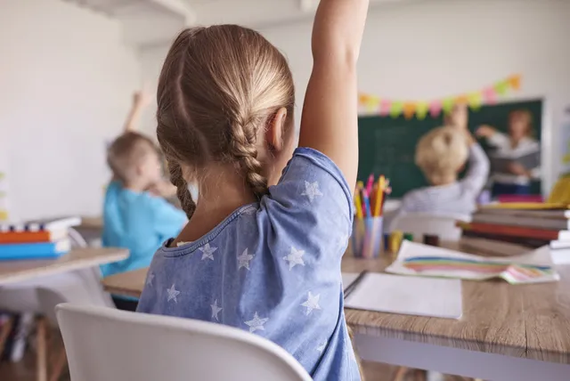 Osztálytermi jelenet: egy fonott hajú kislány hátulról fotózva jelentkezik felemelt kézzel a padjából, mellette és a háttérben más gyermekek is kezüket emelik, elöl a tábla és a tanár körvonala látszik.